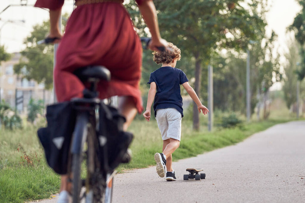 Vista trasera de niño irreconocible en ropa casual montar monopatín en el camino de asfalto en el parque detrás de la mujer de la cosecha en bicicleta - Foto, imagen