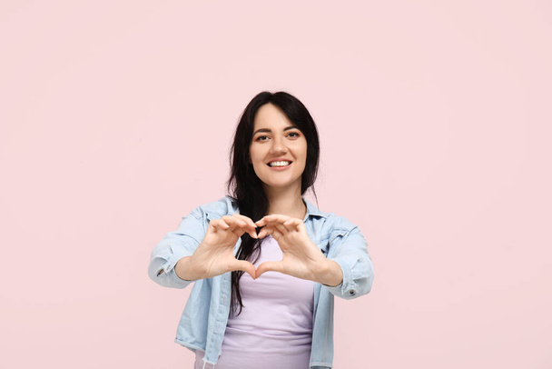 Šťastná mladá žena dělat srdce s rukama na růžovém pozadí - Fotografie, Obrázek