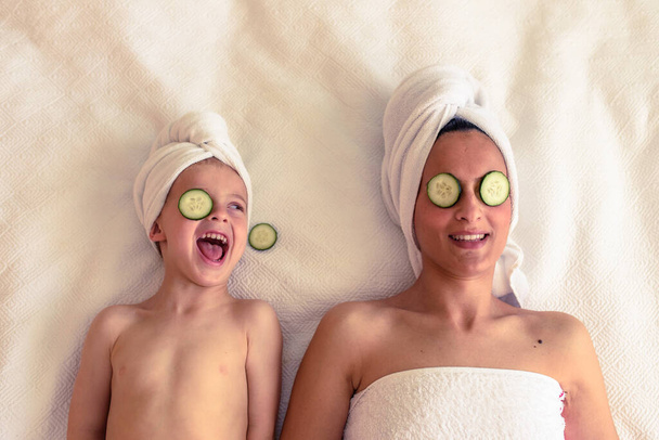 Z góry szczęśliwa kobieta i mały syn relaksujący się na białym kocu z ręcznikami na głowach podczas sesji pielęgnacji skóry z plasterkami ogórka na oczach - Zdjęcie, obraz