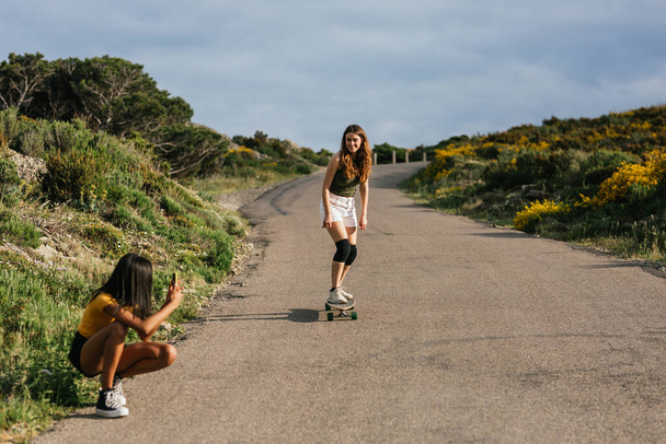Rajattu tunnistamaton etninen naisten selailu älypuhelimella istuessaan longboard asfalttitiellä - Valokuva, kuva