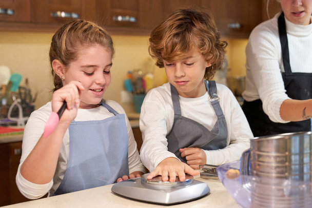 Очаровательные младшие брат и сестра с светлыми волосами и фартуками улыбаются и играют на кухонном весе, помогая матери урожая печь выпечку дома - Фото, изображение
