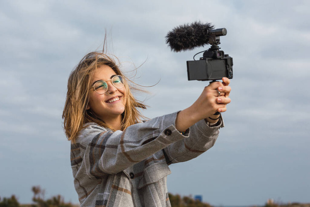 Веселая молодая женщина-путешественница с светлыми волосами в повседневной одежде и очках делает селфи на современной цифровой камере - Фото, изображение