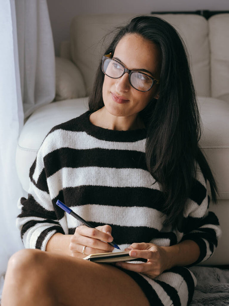 Attractive female in eyeglasses looking into distance with dreamy look and taking notes in notepad while sitting near sofa in light living room - Photo, Image