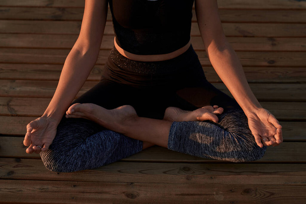 De cima de colheita anônima fêmea descalça em sportswear sentado em pose de lótus com as mãos mudra durante sessão de ioga no caminho de madeira na natureza - Foto, Imagem