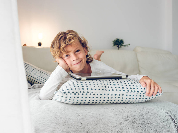 Adorable curly preteen boy lying on comfortable sofa with tablet placed on cushion and looking at camera and smiling slyly - Photo, Image