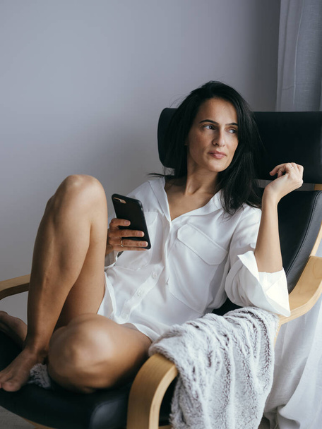 Спокійна молода босоніж етнічна жінка з довгим темним волоссям у білій сорочці мрійливо дивиться, використовуючи мобільний телефон і відпочиваючи в кріслі вдома - Фото, зображення