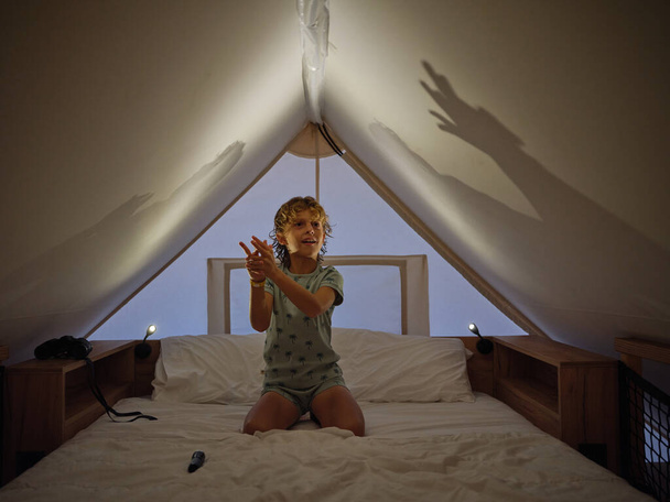 Söpö poika yöpuvussa tehdä varjoja kattoon kädet kun istuu mukava valkoinen sänky illalla kotona - Valokuva, kuva