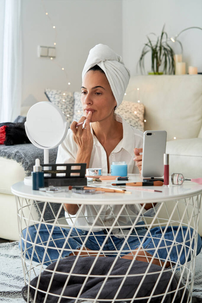 Mujer enfocada con toalla en la cabeza que aplica lápiz labial con el dedo delante del espejo mientras hace maquillaje en casa cerca de la mesa redonda con teléfono inteligente en la mañana - Foto, Imagen