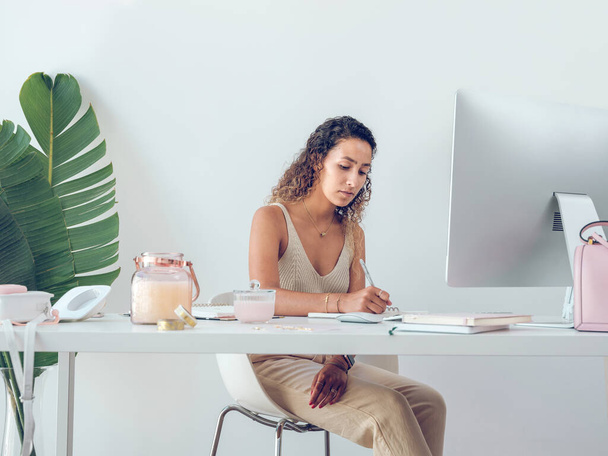Εστιασμένη γυναίκα επιχειρηματίας με σγουρά μαλλιά κάθεται στο τραπέζι με τον υπολογιστή και γράφοντας σημειώσεις στο σημειωματάριο στο φως δωμάτιο - Φωτογραφία, εικόνα