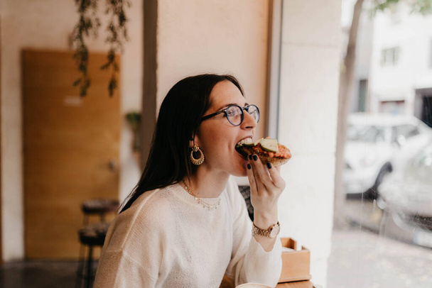 Вид збоку красива молода жінка в стильному вбранні кусає смачний бутерброд, сидячи біля вікна в затишному кафе - Фото, зображення