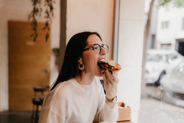 Вид збоку красива молода жінка в стильному вбранні кусає смачний бутерброд, сидячи біля вікна в затишному кафе - Фото, зображення