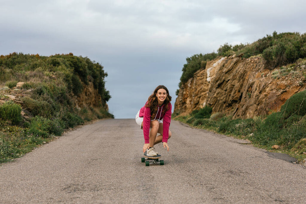 Volledig lichaam van gelukkige jonge vrouw in knie pads rijden longboard op asfalt weg tussen heuvels onder bewolkte hemel - Foto, afbeelding