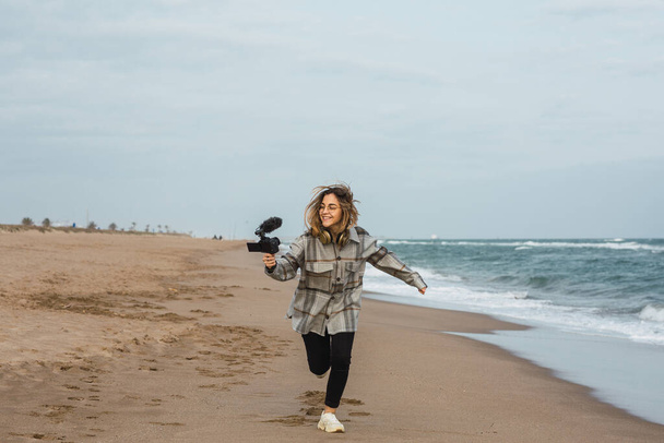 Koko kehon iloinen nuori nainen bloggaaja lämmin ruudullinen paita hymyillen ja käynnissä pitkin hiekkaranta samalla tallennus vlog kameran vasten pilvinen taivas - Valokuva, kuva