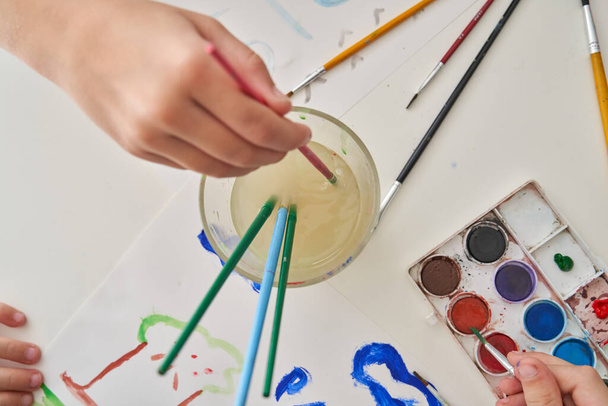 Κάτοψη αγνώριστων ανθρώπων που πλένουν πολύχρωμες βούρτσες και ζωγραφίζουν εικόνες σε λευκά φύλλα χαρτιού χρησιμοποιώντας ακουαρέλες - Φωτογραφία, εικόνα
