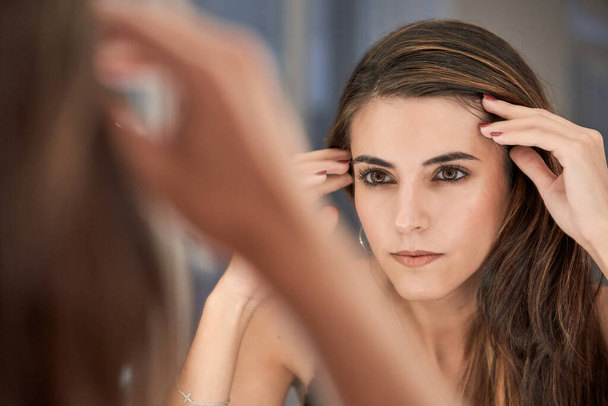 Charmant modèle jeune femme avec maquillage touchant la tête et regardant reflet dans le miroir tout en étant assis dans un salon de beauté moderne - Photo, image