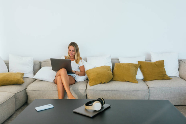 Lächelnde Frau in Freizeitkleidung tippt auf Netbook, während sie auf bequemem Sofa mit Kissen im hellen Wohnzimmer zu Hause sitzt - Foto, Bild