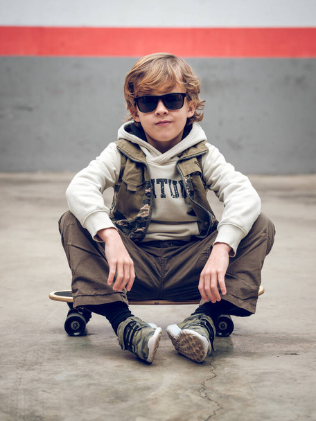 Celé tělo seriózní chlapec ve stylovém oblečení a sluneční brýle sedí na skateboardu na betonové podlaze při pohledu do kamery - Fotografie, Obrázek