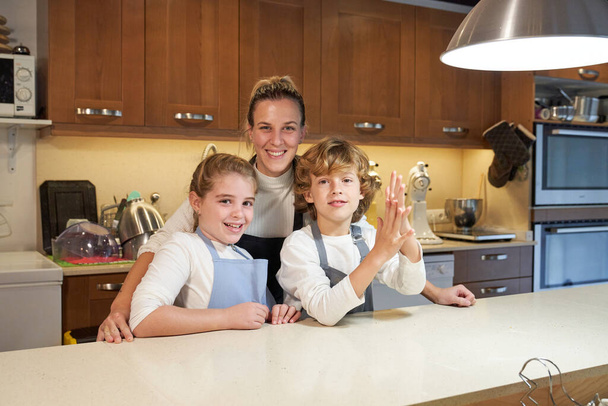 Mosolygó nő és gyerekek kötényt visel áll a pult mellett konyhai eszközök hétvégén, és nézi a kamerát - Fotó, kép