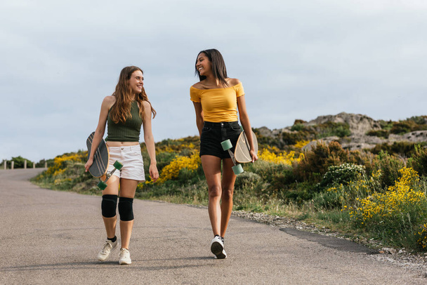Полное тело счастливых многонациональных девушек, смотрящих друг на друга во время прогулки по дороге с длинными досками - Фото, изображение