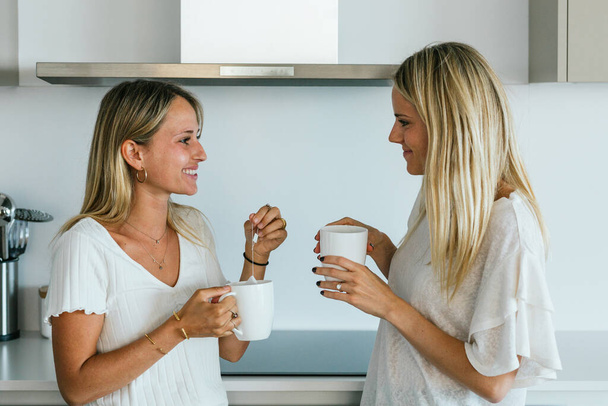 Позитивные подруги с чашками кофе смотрят друг на друга, стоя на кухне с современной посудой дома - Фото, изображение