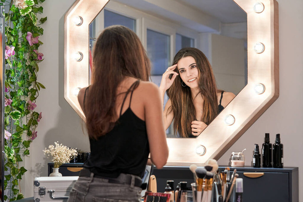 Joyeux jeune femme debout près de la table avec des cosmétiques décoratifs et regarder dans le miroir tout en passant du temps dans un salon de beauté - Photo, image