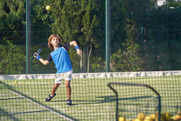 Pełne ciało sportowego chłopca w stroju aktywnym grającego w padel z rakietką na boisku sportowym z siatką podczas treningu w letni dzień - Zdjęcie, obraz