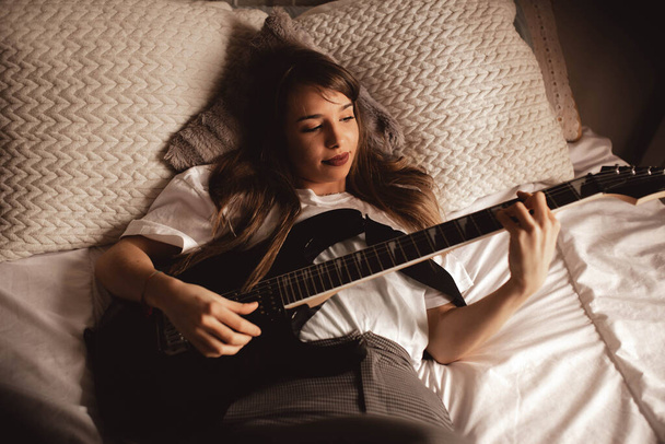 上から若いブルネットの女性がベッドの上でギターを弾く - 写真・画像