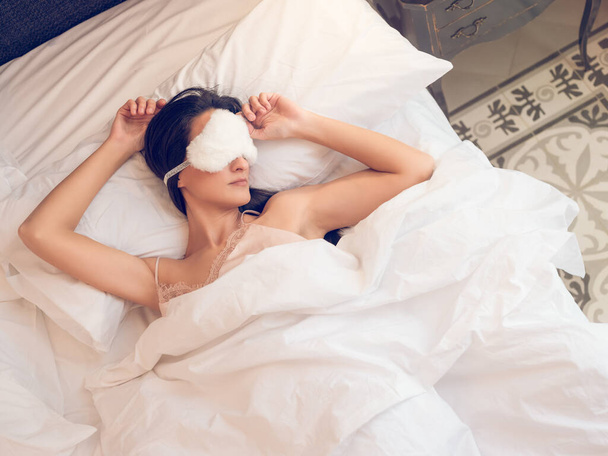Widok z góry młoda kobieta z czarnymi włosami i w śpiochach relaks na wygodnym łóżku z maską na twarzy i z podniesionymi ramionami na poduszce rano - Zdjęcie, obraz