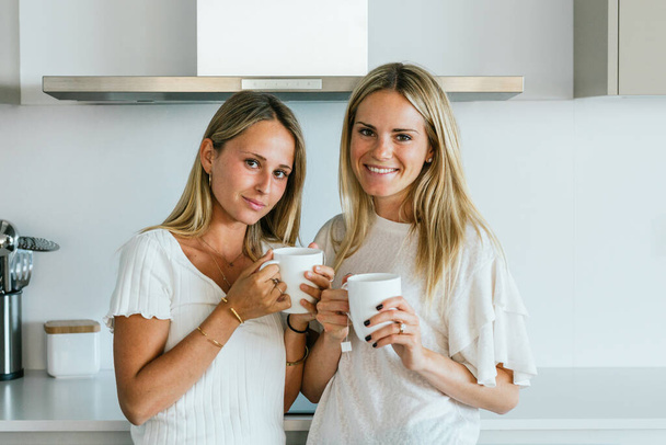 Positive female friends with cups of coffee looking at camera while standing in kitchen with modern kitchenware at home - Photo, Image