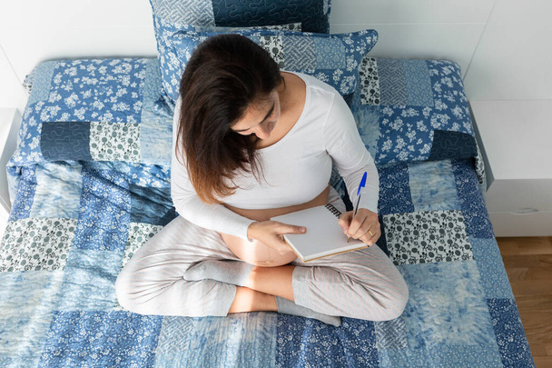 From above of pregnant female sitting on bed and writing in notebook in morning - 写真・画像
