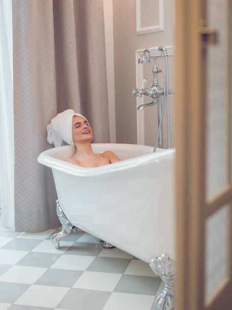 Jonge vrouw met handdoek op het hoofd ontspannen met gesloten ogen in bad, terwijl glimlachen in de moderne badkamer - Foto, afbeelding