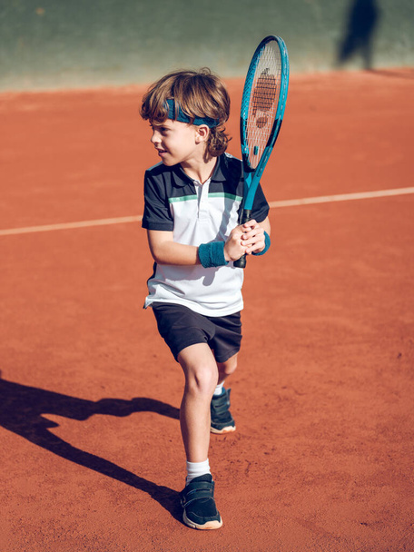 Cuerpo completo de adorable niño preadolescente enfocado en ropa deportiva preparándose para el derrame cerebral con raqueta mientras juega al tenis en pista dura - Foto, Imagen
