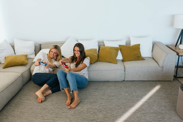 Corpo inteiro de amigas positivas com controladores de jogos jogando videogame enquanto sentadas no chão apoiadas no sofá na sala de estar em casa - Foto, Imagem