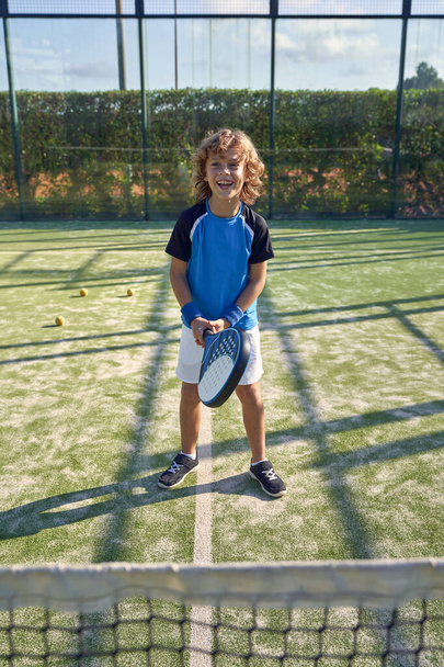 Pełne ciało pozytywnego chłopca w odzieży sportowej z rakietą odwracając wzrok stojąc na trawiastym placu zabaw z siatką podczas gry padel w letni dzień - Zdjęcie, obraz