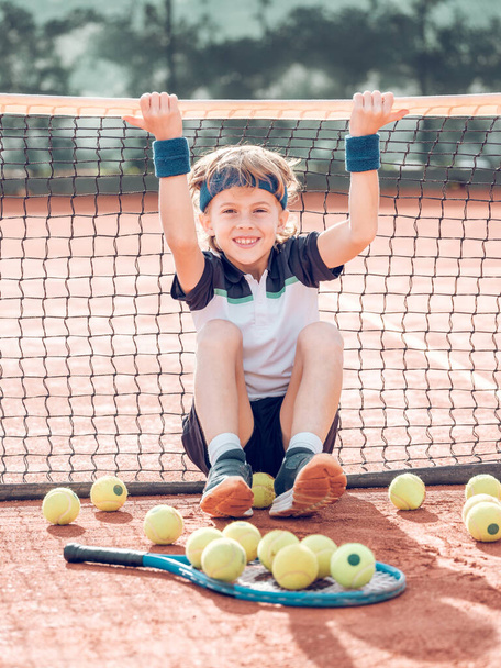 Pełne ciało pozytywnego chłopca z blond włosami w stroju aktywnym i tenisówkach uśmiechającego się i patrzącego w kamerę, opierającego się na siatce siedzącej na korcie glinianym podczas treningu tenisowego - Zdjęcie, obraz