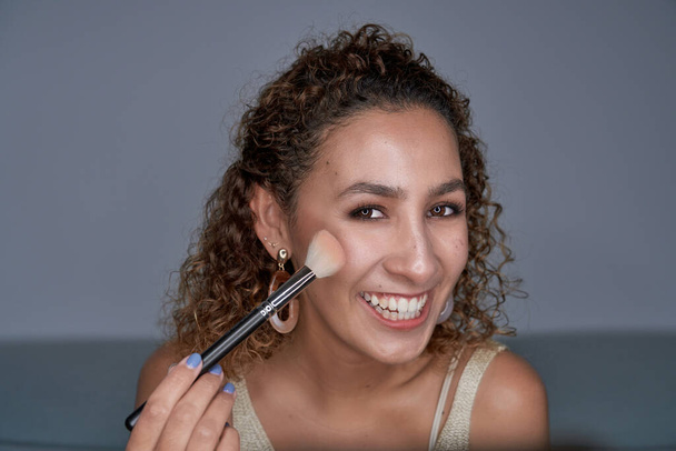 Nadšené kudrnaté vlasy ženské visagistky nanášení zářící zvýrazňovač prášek na tvář s profesionálním štětcem a při pohledu na fotoaparát - Fotografie, Obrázek