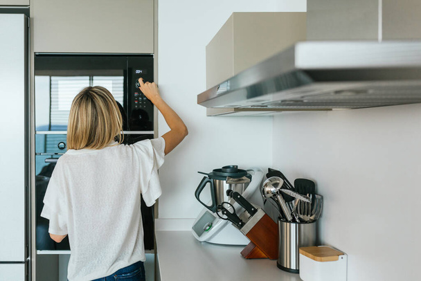 Vista posteriore di anonima femmina in abito casual accendere moderno forno a microonde nero mentre in piedi in cucina con utensili da cucina moderni - Foto, immagini