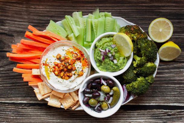 From above appetizing healthy hummus green sauce cut carrot cucumbers and green broccoli salted cucumbers decorated with sliced lemon in white bowls on wooden table - Photo, Image