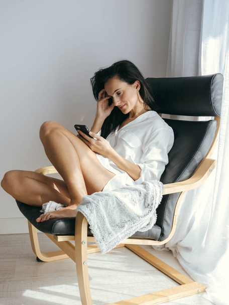 Widok na całe ciało uroczej kobiety z gołymi nogami SMS-y na komórce siedząc w fotelu przy oknie - Zdjęcie, obraz