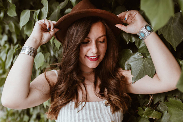 Çekici, uzun saçlı, yeşil yaprakların ortasında şapkalı mutlu kadın. - Fotoğraf, Görsel