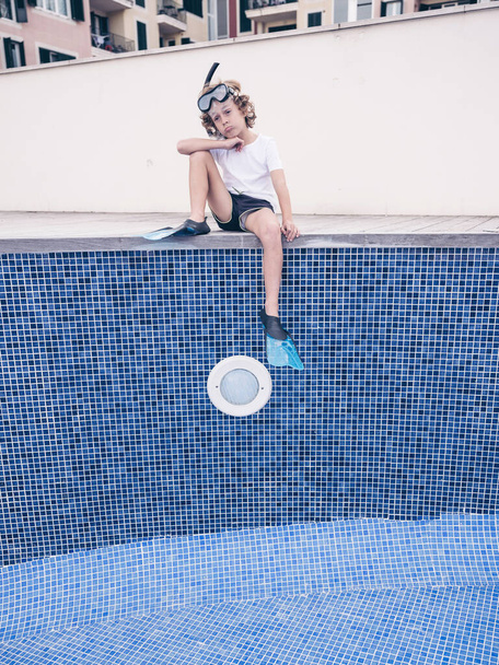 Desde abajo de cuerpo completo preguntándose niño preadolescente en máscara de snorkel y aletas sentado con la pierna doblada en el lado de la piscina vacía - Foto, imagen