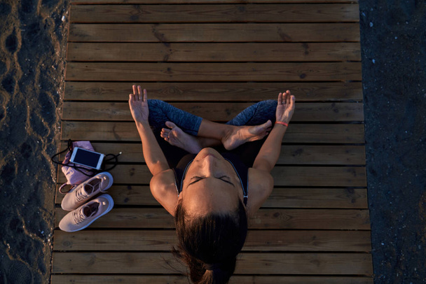 Top view full body of barefoot female meditating in lotus pose with mudra hands while sitting on boardwalk on shore - Φωτογραφία, εικόνα