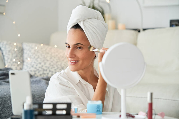 Positive adult smiling female with towel on head applying liquid foundation with brush while shooting video for beauty blog at home sitting near sofa - Photo, Image
