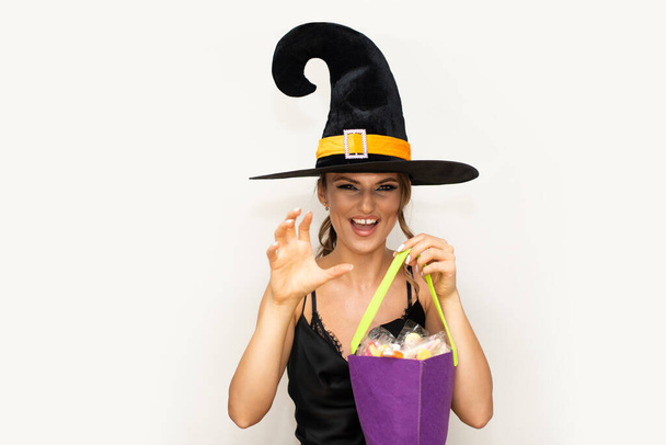Młoda kobieta w czarnym kapeluszu czarownicy w czarnej jedwabnej sukience przeraża się dłonią i zębami, trzymając koszyk pełen słodyczy Halloween i smakołyków - Zdjęcie, obraz