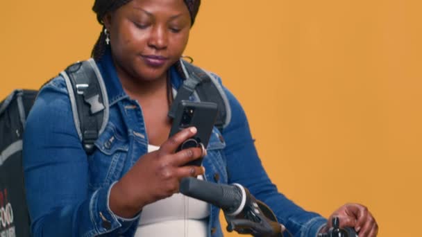 Ενθουσιώδης παράδοση γυναίκα μηνυμάτων συναδέλφους με smartphone στο ποδήλατο πριν από την ποδηλασία για την υπηρεσία παράδοσης τροφίμων. African American courier έλεγχο κινητό app για πακέτα για την παράδοση on-demand. - Πλάνα, βίντεο