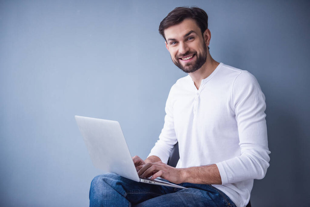 Hombre joven guapo está utilizando un ordenador portátil, mirando a la cámara y sonriendo, sobre fondo gris
 - Foto, Imagen