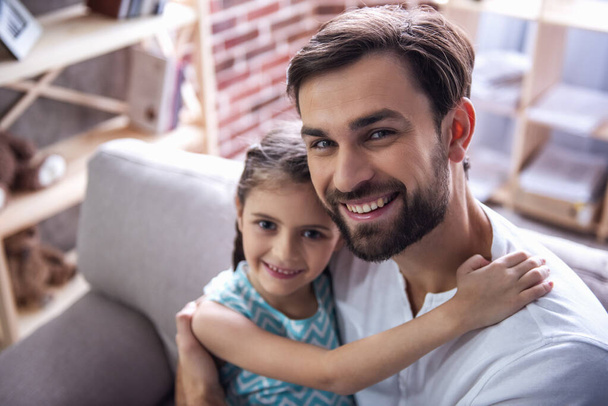 Милая маленькая девочка и ее красивый отец обнимаются, смотрят в камеру и улыбаются, сидя дома на диване
 - Фото, изображение