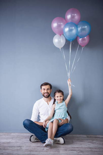 niedliches kleines Mädchen und ihr hübscher Vater halten Luftballons in die Kamera und lächeln auf grauem Hintergrund - Foto, Bild