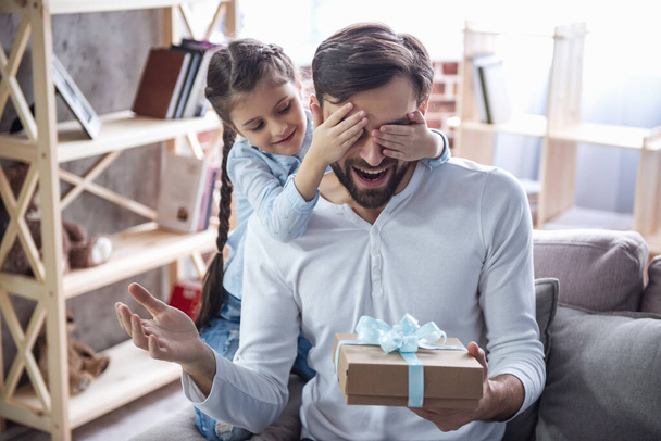 Nettes kleines Mädchen schenkt ihrem gutaussehenden Vater eine Geschenkbox. beide sitzen zu Hause auf der Couch und lächeln - Foto, Bild