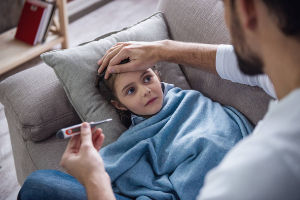 Маленькая больная девочка, покрытая одеялом, лежит на диване, пока её отец измеряет ей температуру.
 - Фото, изображение
